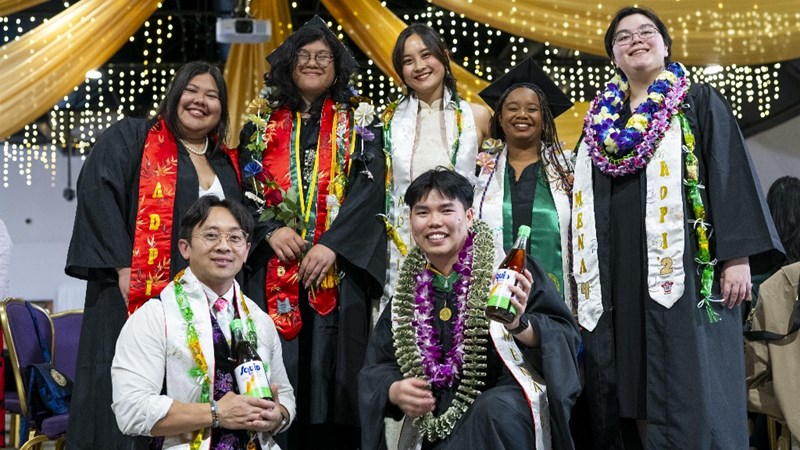 Seven smiling students in graduation regalia with cultural sashes. 