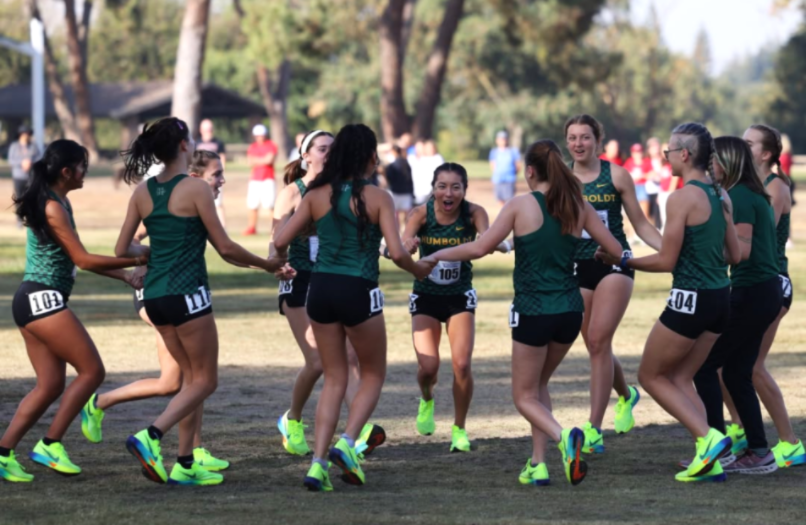 11 women's cross country teammates holding hands in a circle smiling and laughing.