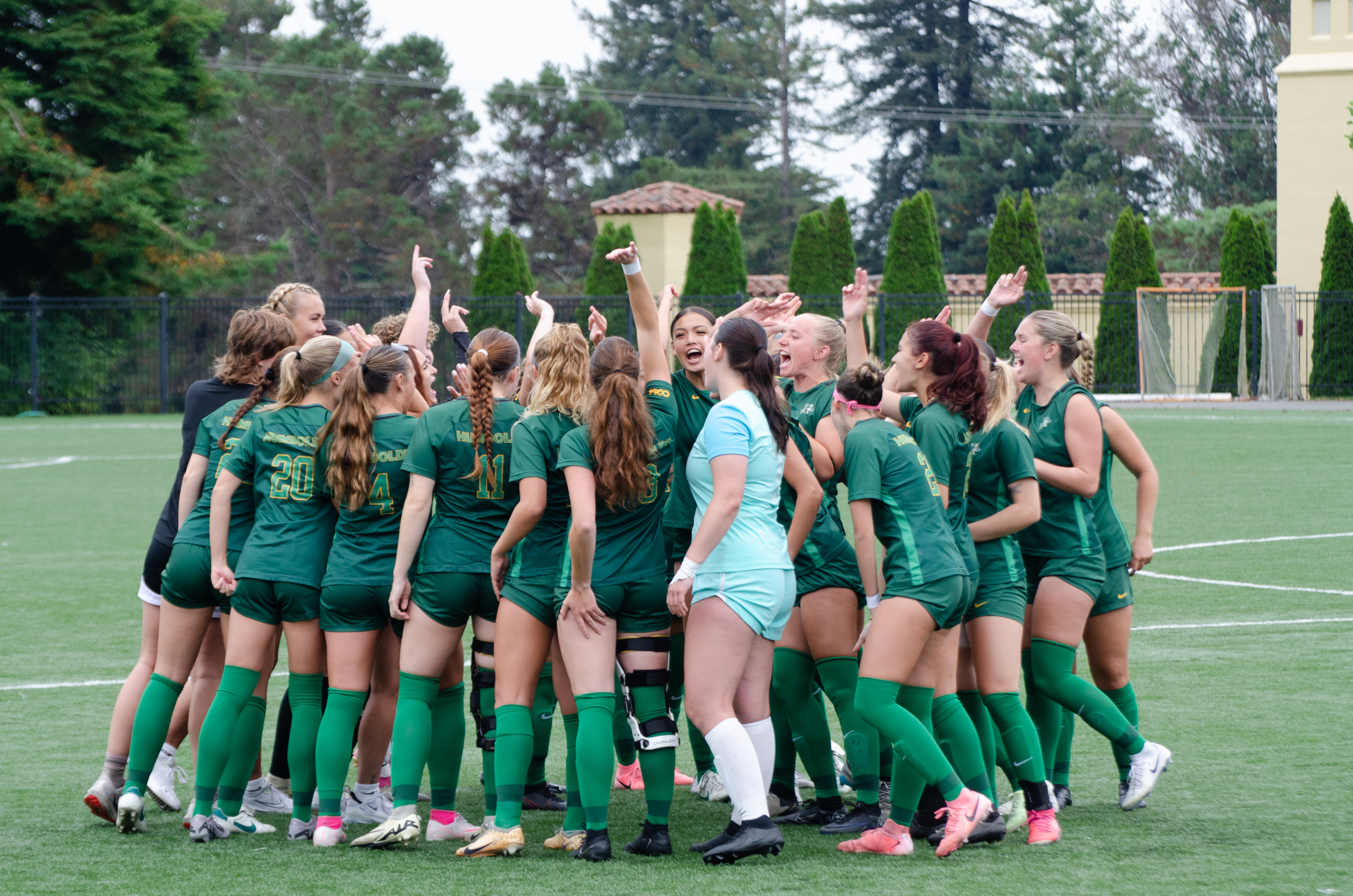 The women's soccer team in green jerseys in a huddle on the soccer field.