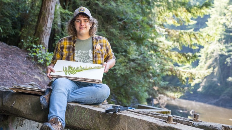 A student sitting by a river and holding a fern in their hand.