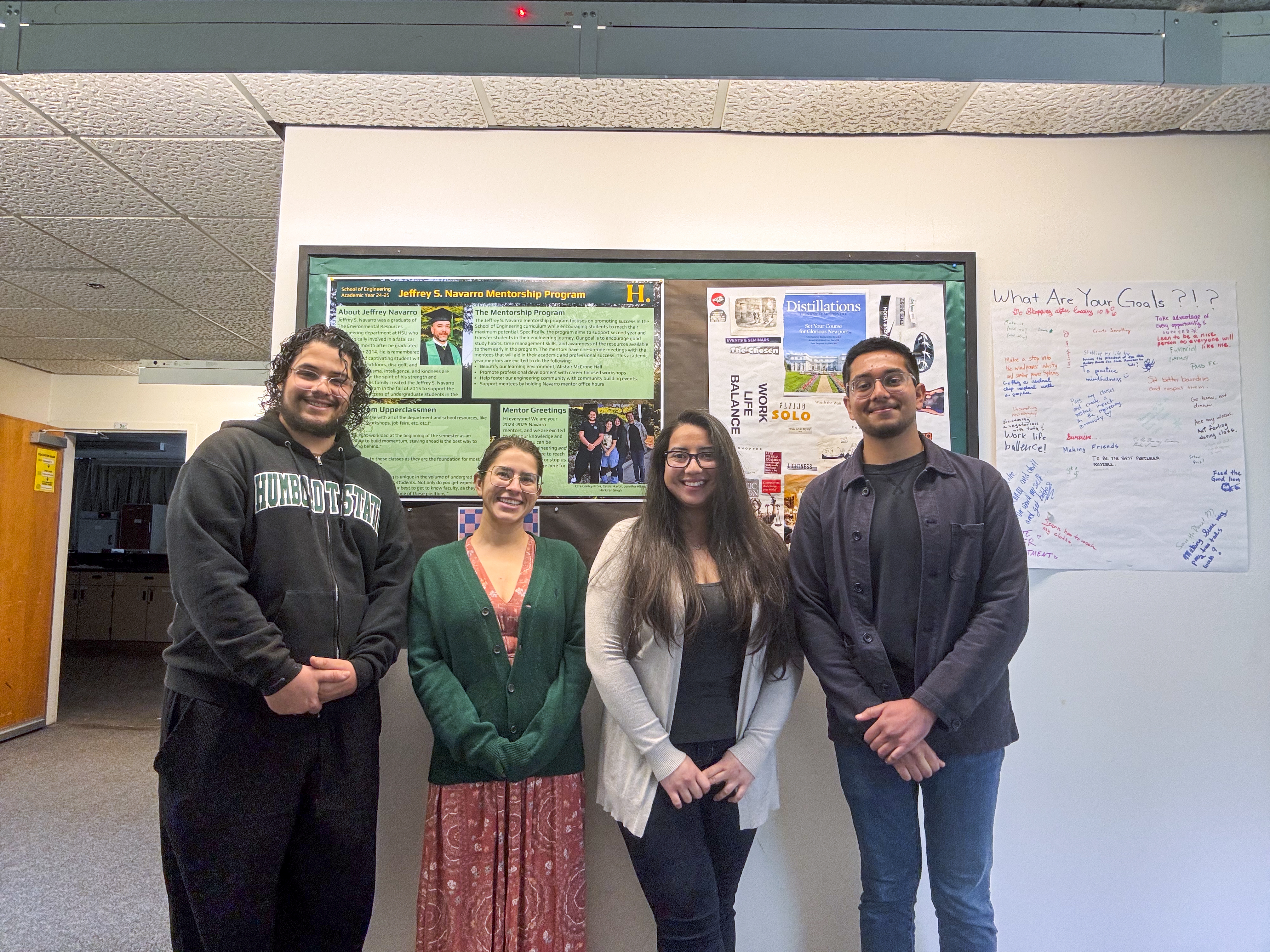 Four people smiling and standing in front of a mentorship program poster.