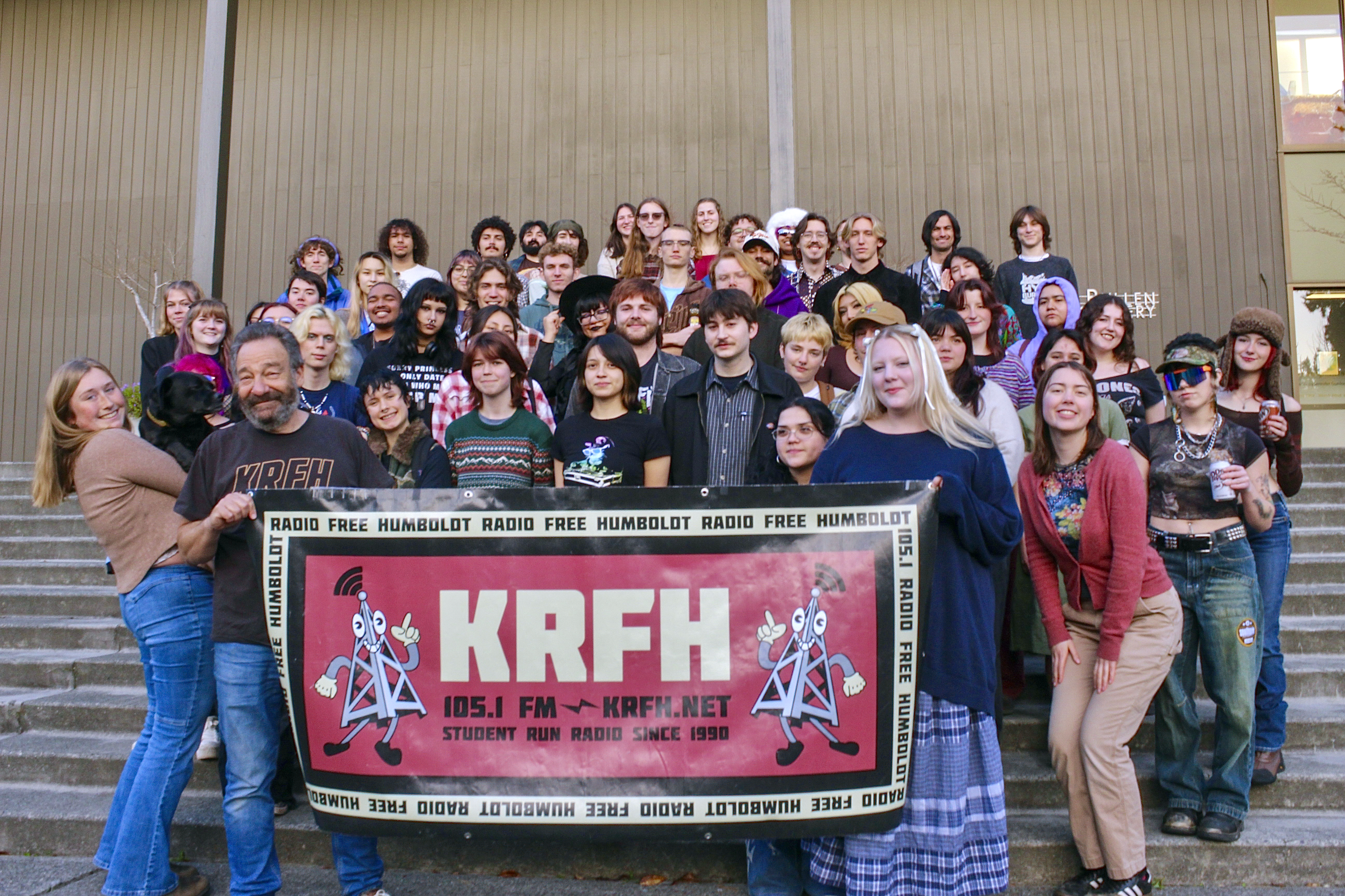 The KRFH student staff and professor holding a KRFH banner in front of the step of the Art building.