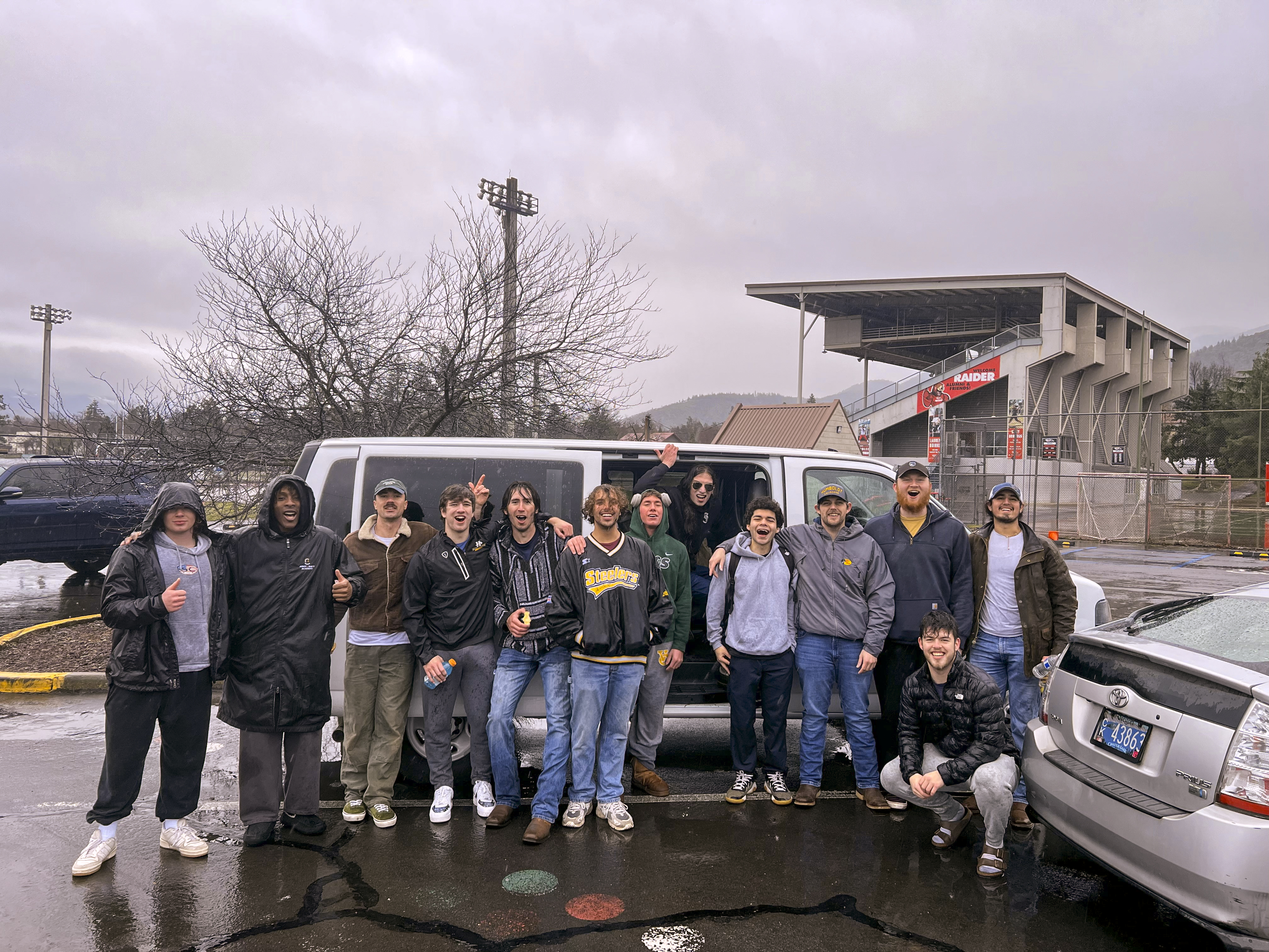 Group photo of men's lacrosse players in the rain outside of a van.