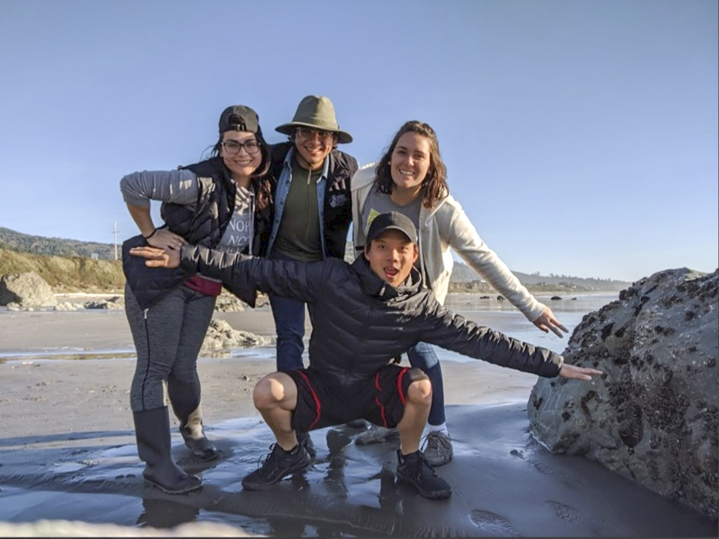 Four students smiling at the beach next to a large rock.