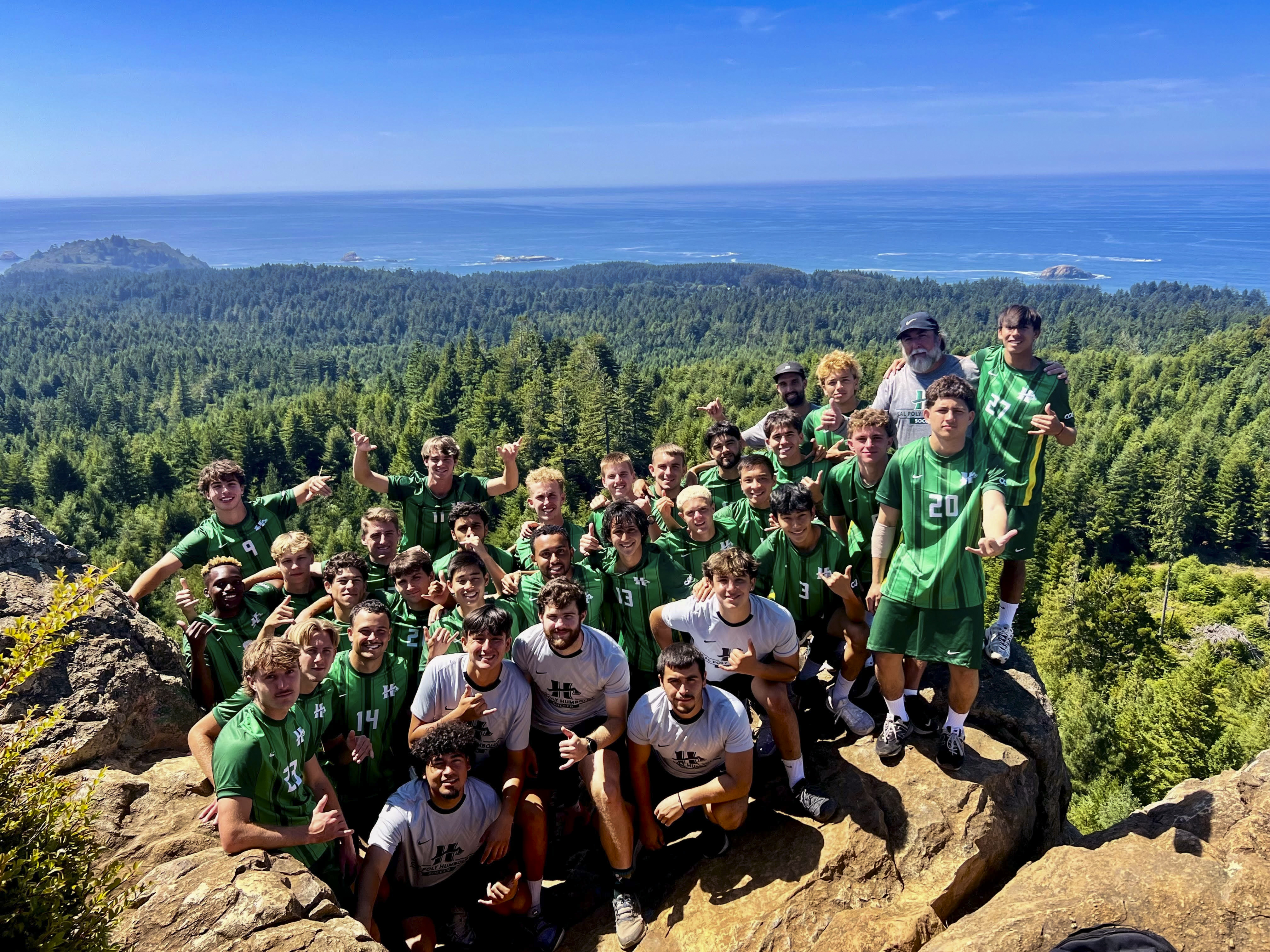 A group photo of the men's soccer team on top of a rock with the ocean in the background.