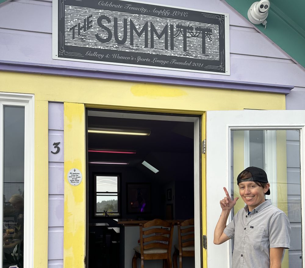 A photo of Michelle Cartier pointing to the sign of The Summitt at the gallery-space's entrance. 