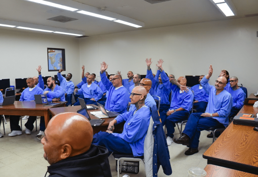 A photo of inmate students, some raising their hand, in class.