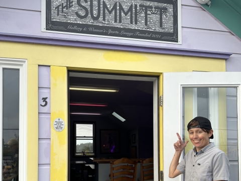 A photo of Michelle Cartier pointing to the sign of The Summitt at the gallery-space's entrance. 