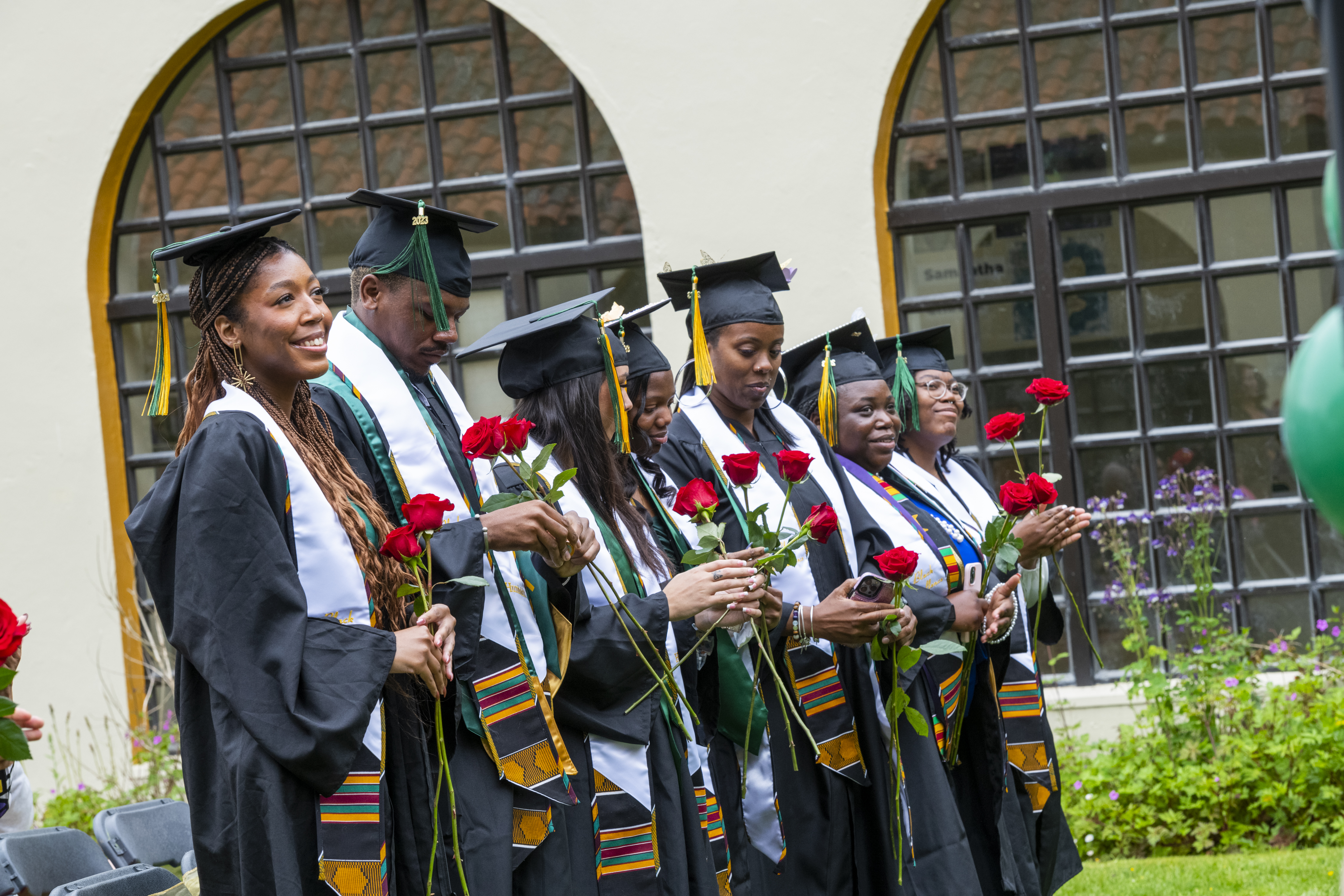 Seven Umoja grads in commencement regalia outside smiling.